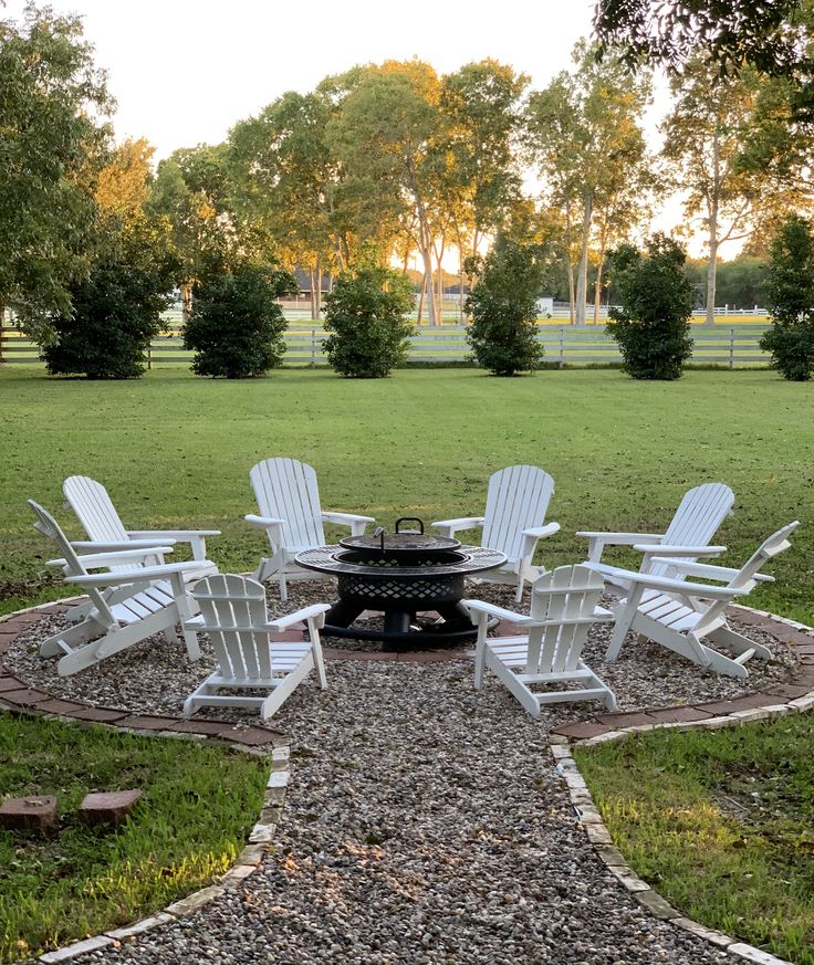 a fire pit surrounded by white chairs in the middle of a grassy area with trees