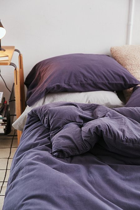 an unmade bed with purple comforter and pillows on the floor next to a lamp