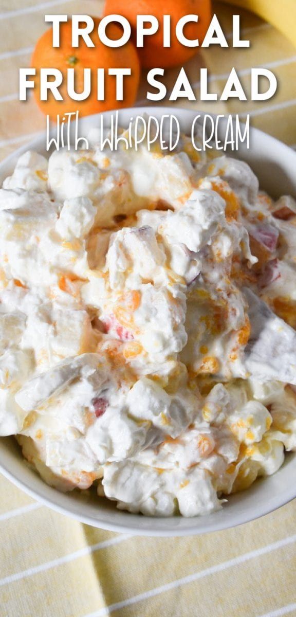 tropical fruit salad with whipped cream in a white bowl on a yellow tablecloth next to an orange