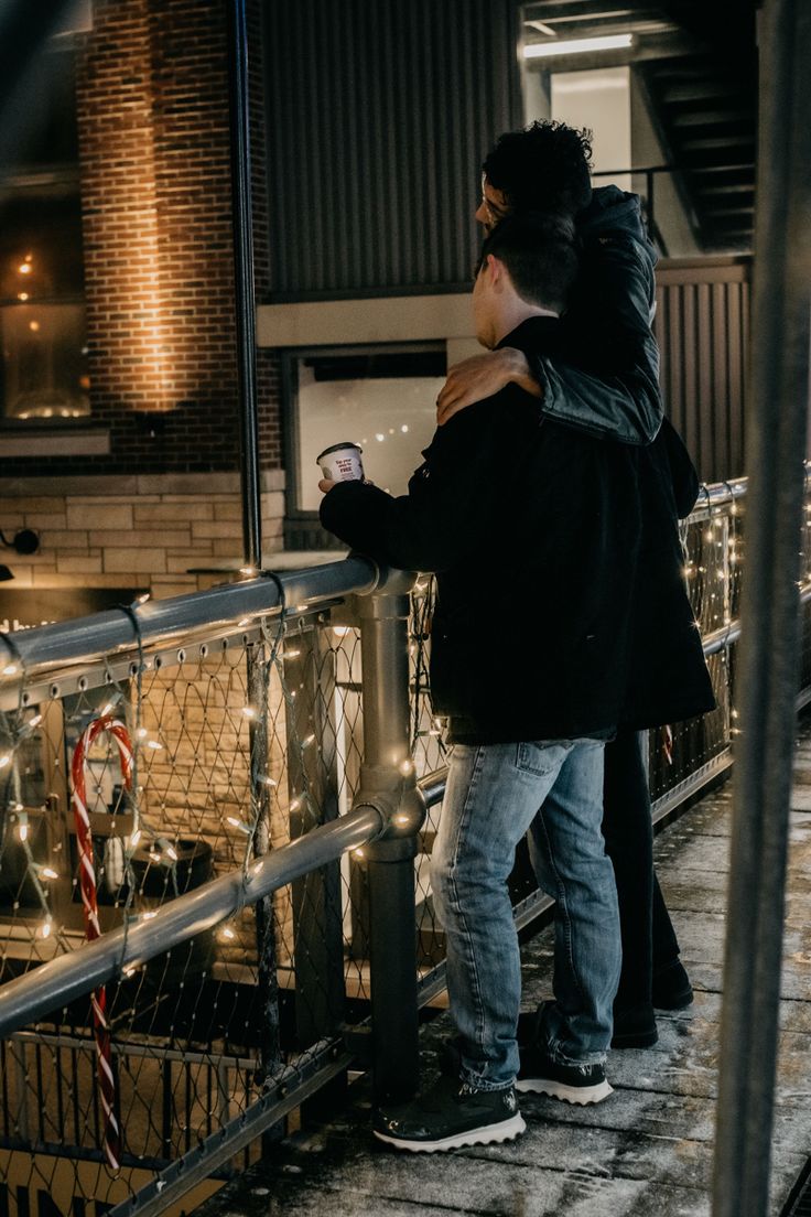 two people standing next to each other near a fence with christmas lights on the railing