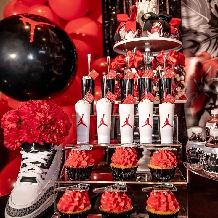 a table topped with cupcakes and cakes covered in red icing next to balloons