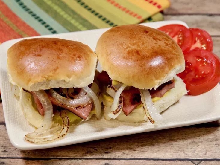 two sandwiches with onions and meat on a plate next to tomato slices, lettuce and tomatoes