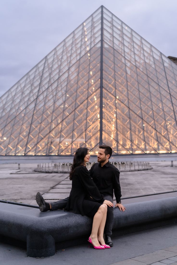 a man and woman sitting on a bench in front of a glass pyramid at night