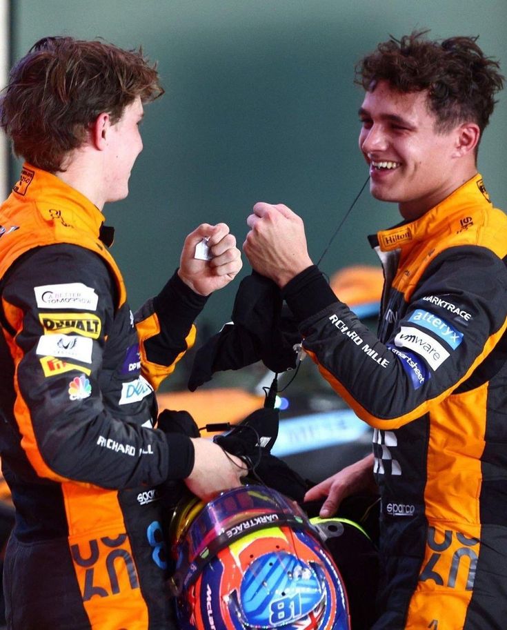 two young men standing next to each other on top of a race track with helmets around their necks