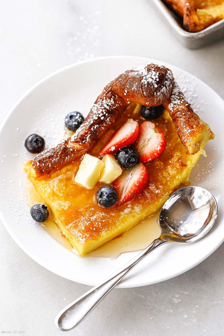 a white plate topped with french toast covered in berries and powdered sugar next to a bowl of fruit