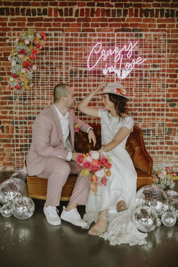 a bride and groom sitting on a bench in front of a brick wall with neon sign