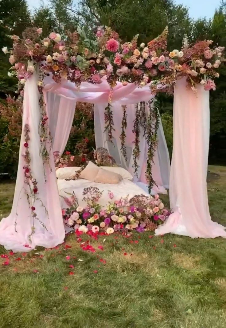 a bed covered in pink flowers and white drapes on top of a lush green field