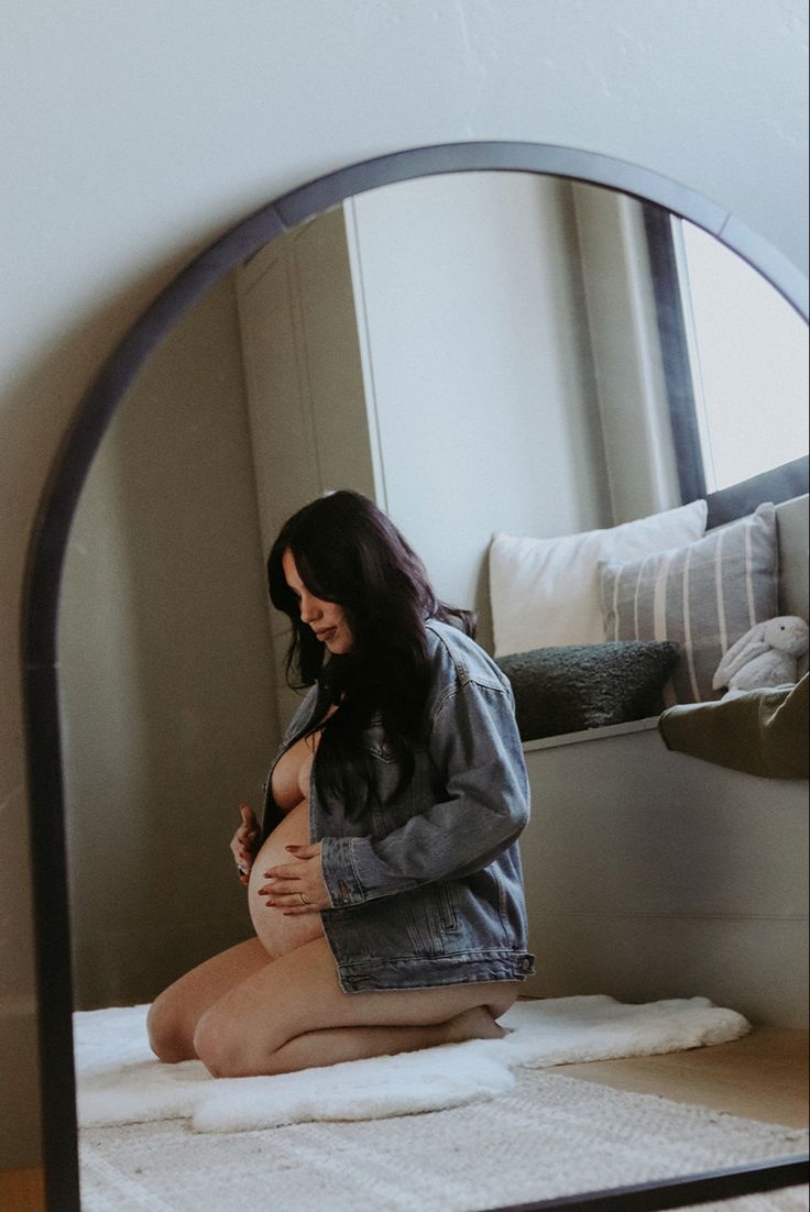 a pregnant woman sitting on top of a bed in front of a large round mirror