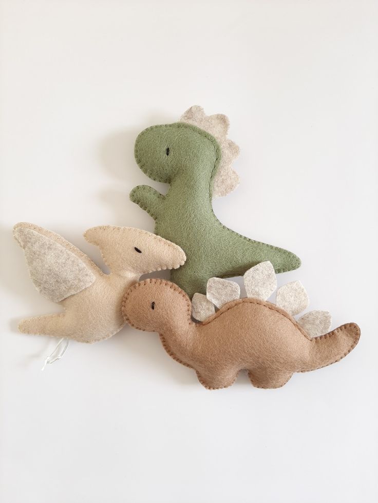 two stuffed dinosaurs sitting next to each other on a white surface, one is green and the other is brown