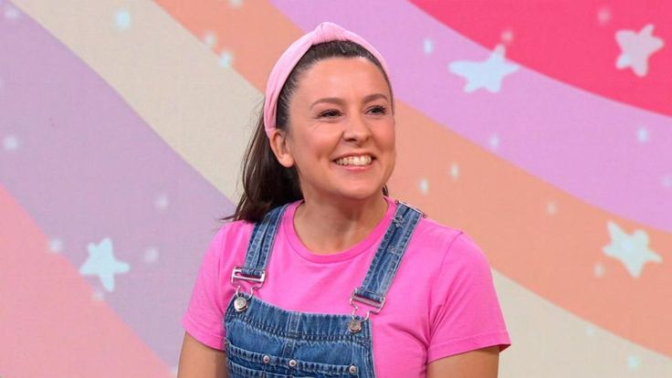 a woman wearing overalls and a pink headband smiles at the camera while standing in front of a colorful background