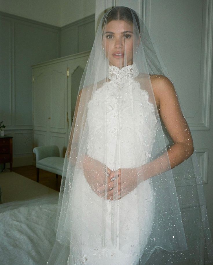 a woman wearing a wedding dress and veil standing in front of a bed with her hands on her hips