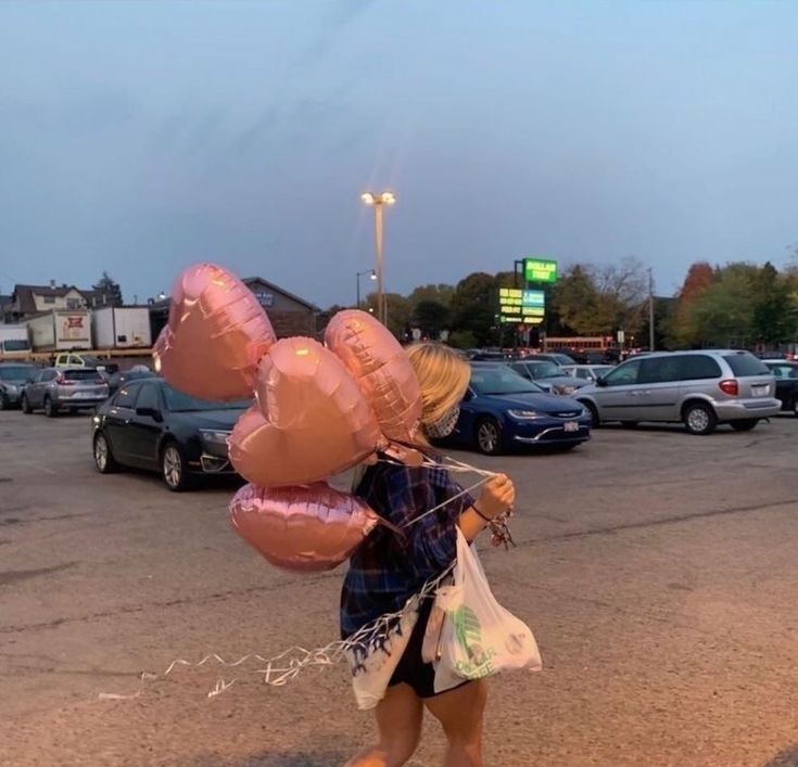 a woman walking across a parking lot holding pink balloons in the shape of heart shapes