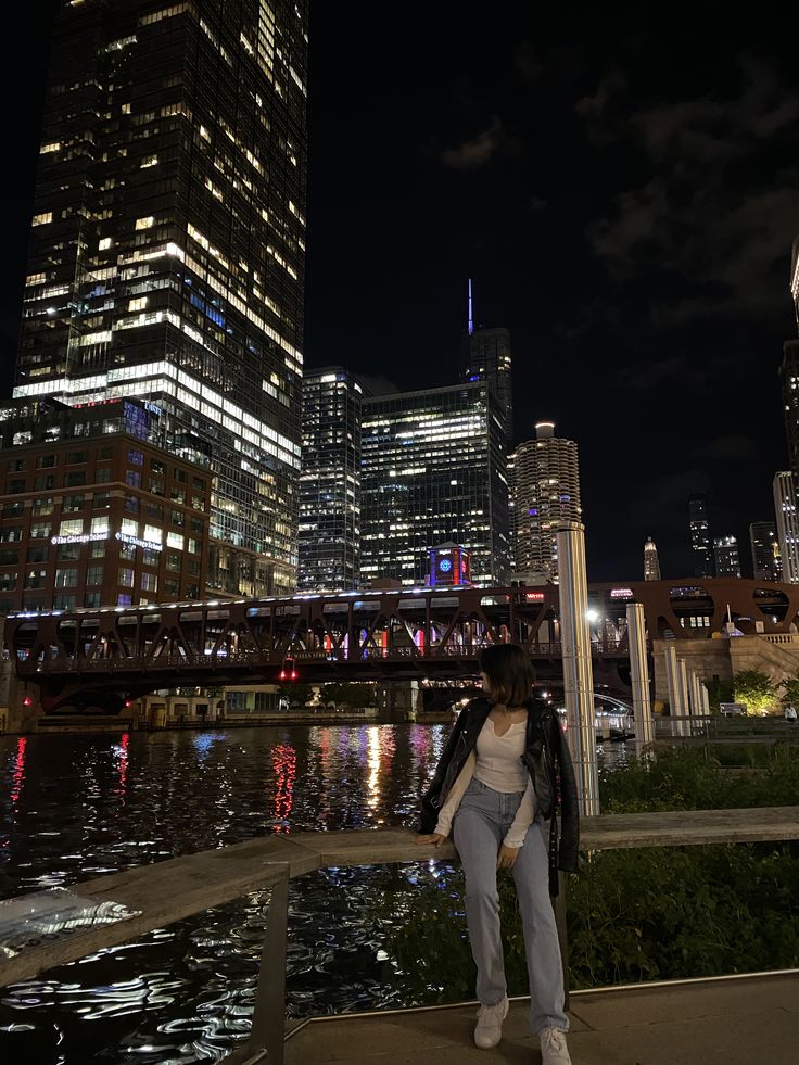 a woman standing on the side of a river in front of tall buildings at night