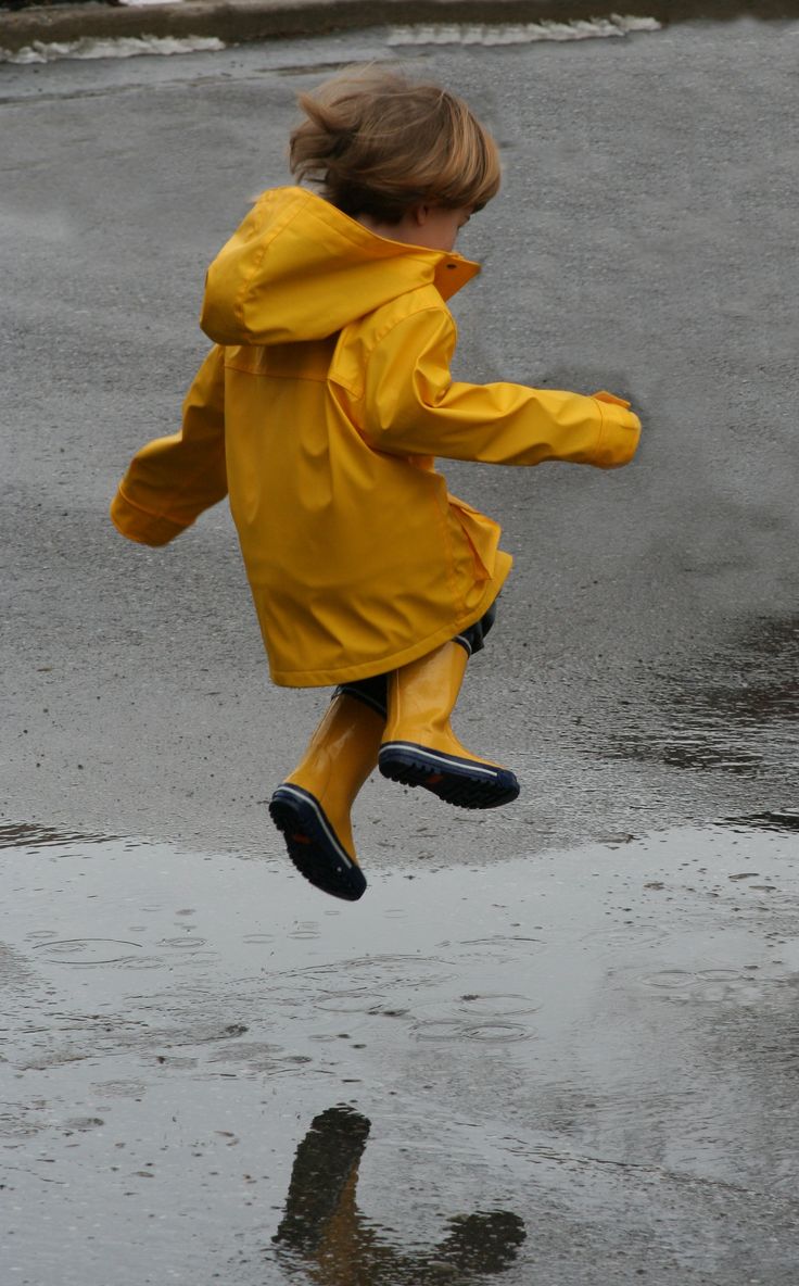 a little boy in a yellow rain coat jumping into the air on a wet sidewalk