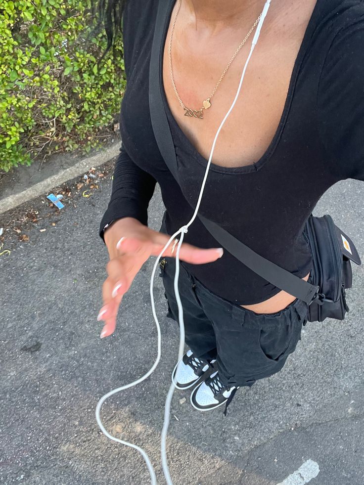 a woman in black shirt and headphones standing on street next to bushes with her hand out