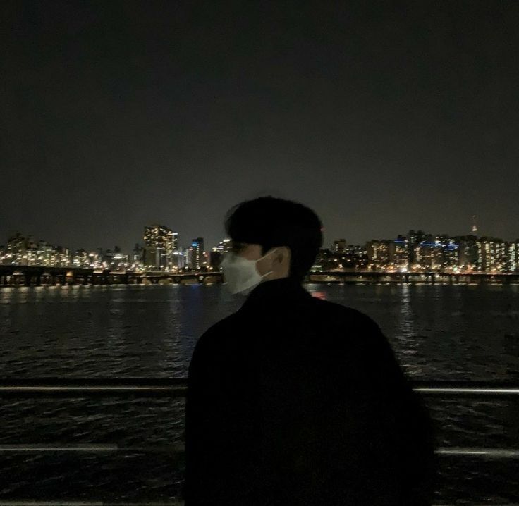 a man wearing a face mask looking out over the water at night with city lights in the background