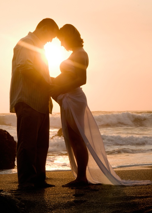 a bride and groom standing on the beach at sunset with their arms around each other