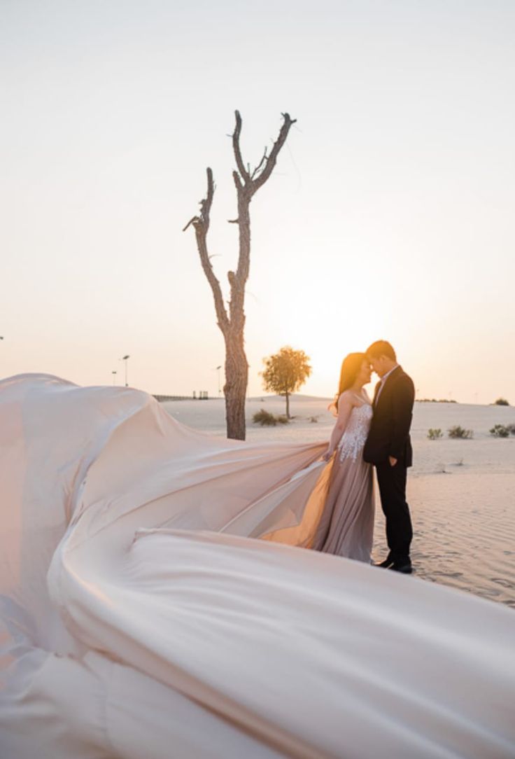 a bride and groom standing in the sand at sunset with their wedding dress blowing in the wind