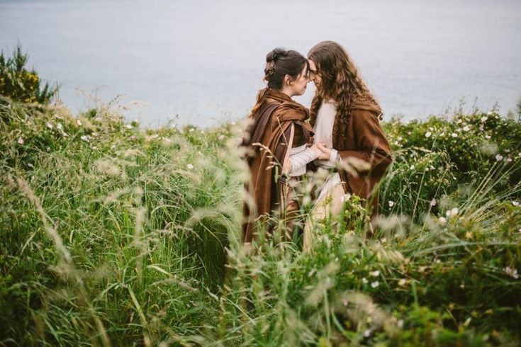 two women are standing in tall grass by the water with their backs to each other