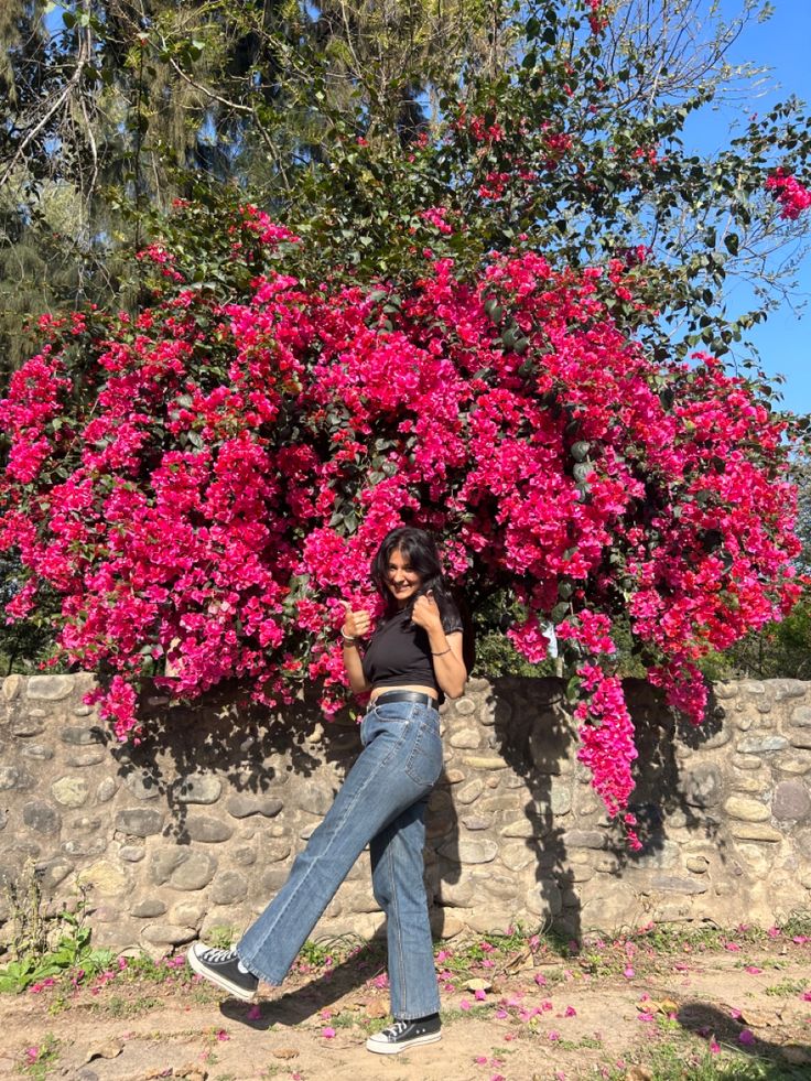 a woman leaning against a stone wall while talking on her cell phone in front of a flowering tree