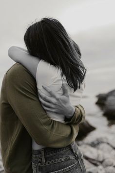 two people embracing each other on rocks near the water with their backs to one another