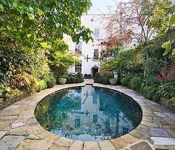 an outdoor swimming pool surrounded by greenery and stone walkways in front of a white house