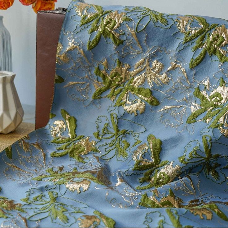 a blue couch covered in green and gold leafy fabric next to a vase with orange flowers