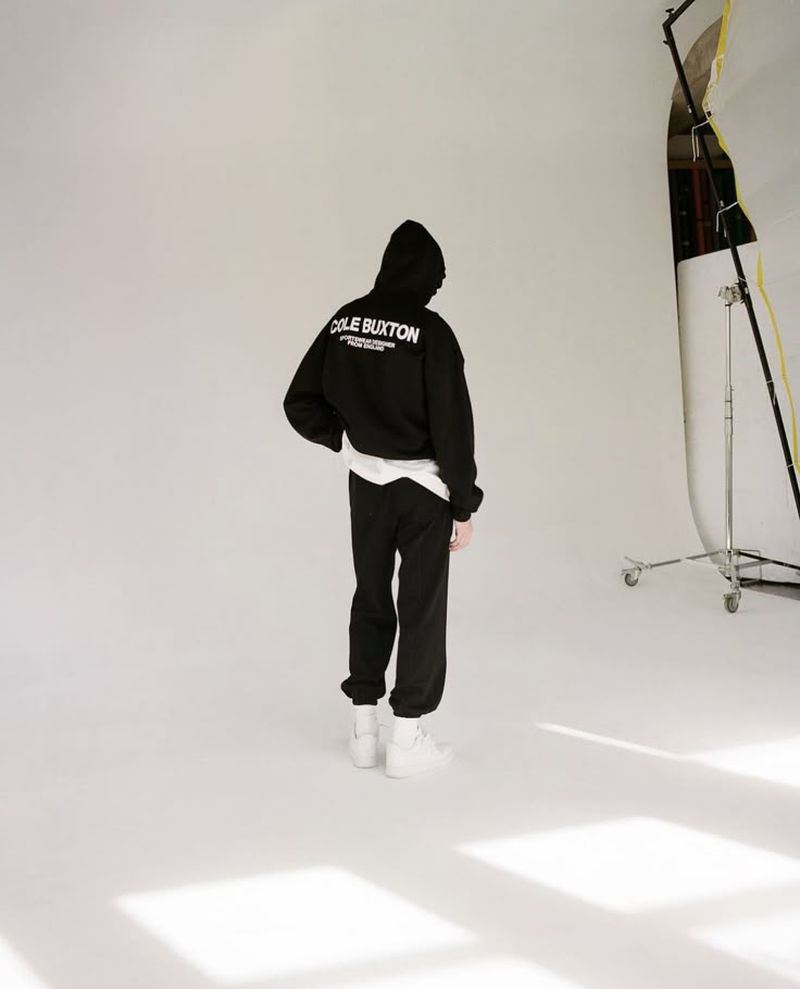 a person standing in front of a white backdrop wearing a black sweatshirt and sweatpants