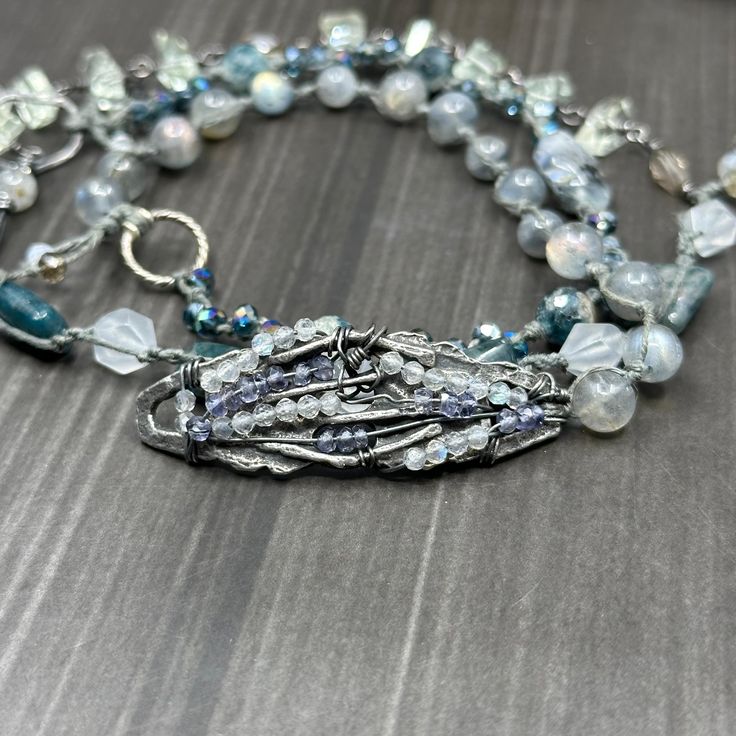 This incredible wrap bracelet has been a work in progress for a while. The combination of the pewter focal with the gorgeous 6mm flashy labradorite rounds absolutely enchanted me. From there, I added so many of my favorite things. In this bracelet, you’ll find apatite rectangles, the most insane faceted 8mm kyanite, 8mm starcut pale jade, flashy tourmilinated rainbow moonstone, smoky quartz, and the most incredible faceted prasiolite nuggets. Also featured are the prettiest faceted glass rondelles. Let’s circle back to the focal. The shape was so interesting and mysterious to me. I wrapped on faceted labradorite and iolite to bring out some of the lines. This wrap bracelet is sterling silver, with the exception of the USA made pewter focal. The knotted portion of the bracelet was done in h Artisan Silver Beaded Bracelets With Unique Variations, Silver Multi-strand Hand Wrapped Bracelets, Silver Hand Wrapped Beaded Bracelets For Healing, Silver Gemstone Beads Wrap Bracelet, Silver Multi-strand Spiritual Bracelets, Silver Gemstone Beads Wrap Bracelet As Gift, Silver Double Strand Bracelet With Natural Stones, Silver Double Strand Beaded Bracelets With Gemstones, Silver Wrap Bracelet With Gemstone Beads