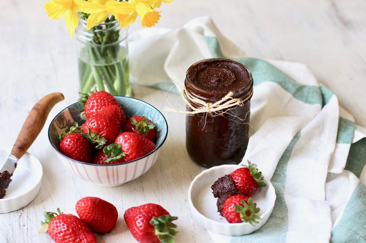 some strawberries are in small bowls next to a jar of chocolate butter and flowers