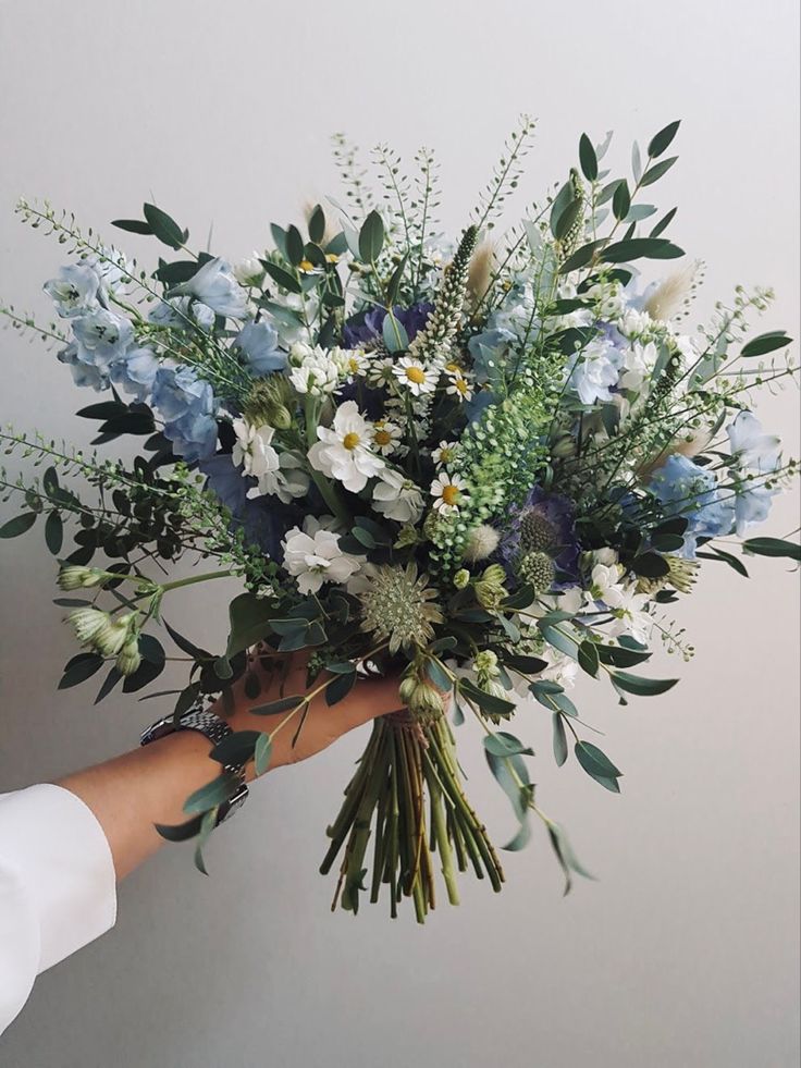 a person holding a bouquet of blue and white flowers with greenery in their hand