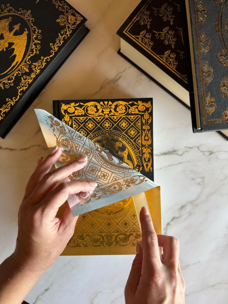 a person holding an envelope in front of two books on a table with gold and black designs