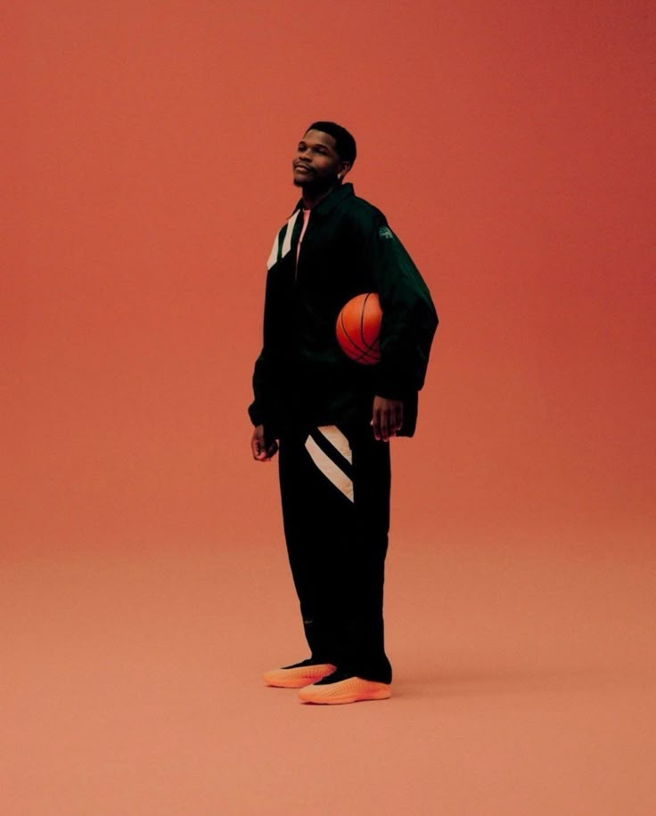 a man standing in front of a red background with a basketball on his foot and wearing a black sweat suit