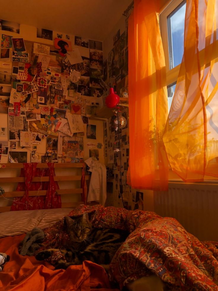 an unmade bed sitting in front of a window covered with pictures and orange curtains