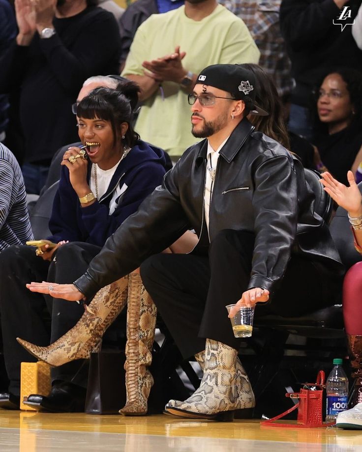 a man in black jacket and snake skin boots sitting on the sidelines at a basketball game