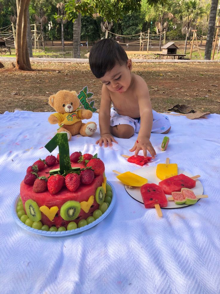 a little boy that is sitting in front of a cake with fruit on it and a teddy bear