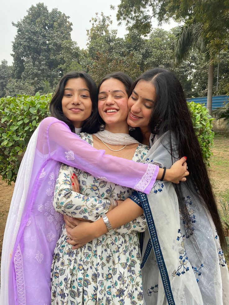 three young women hugging each other in front of trees and bushes, one wearing a purple scarf