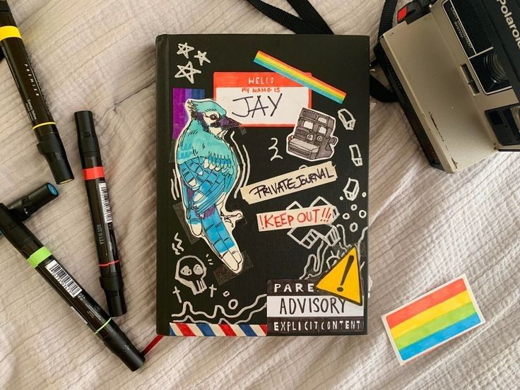 an open notebook sitting on top of a bed next to some markers and a camera