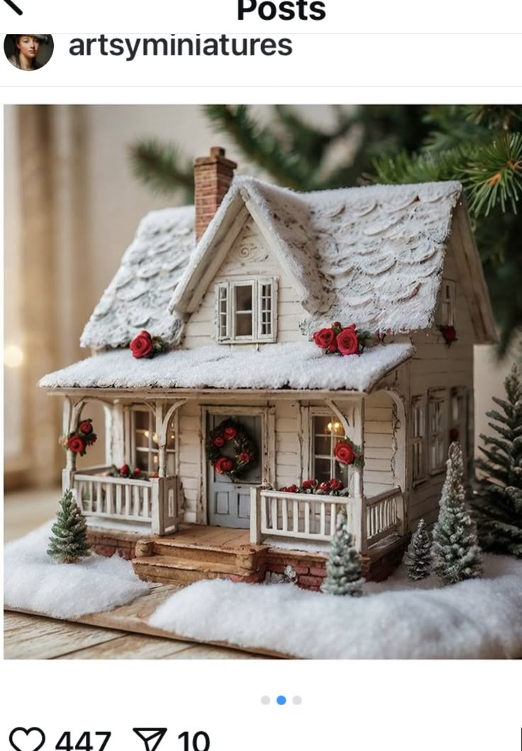 a small white house with snow on the roof and trees around it, in front of a christmas tree