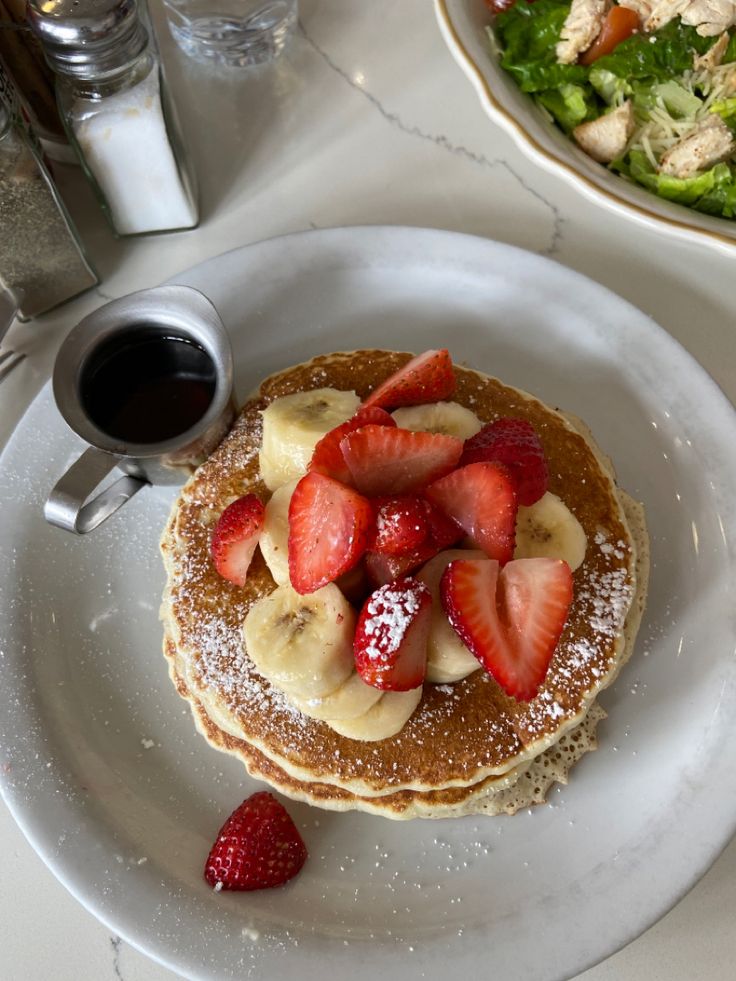 a white plate topped with pancakes covered in bananas and strawberries next to a bowl of salad