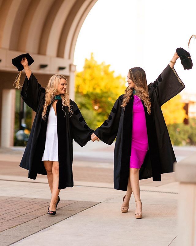 two women in graduation gowns hold hands as they walk down the street with their arms in the air