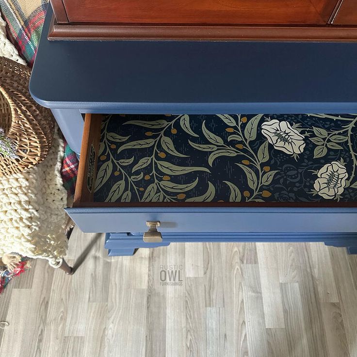 a blue table with a floral design on the top and bottom drawer, next to a wicker basket