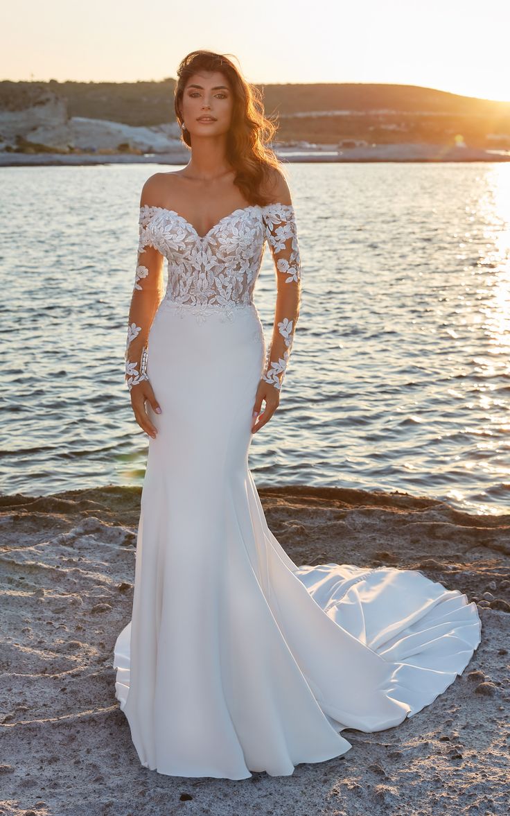 a woman in a white wedding dress standing on the beach with her arms behind her back