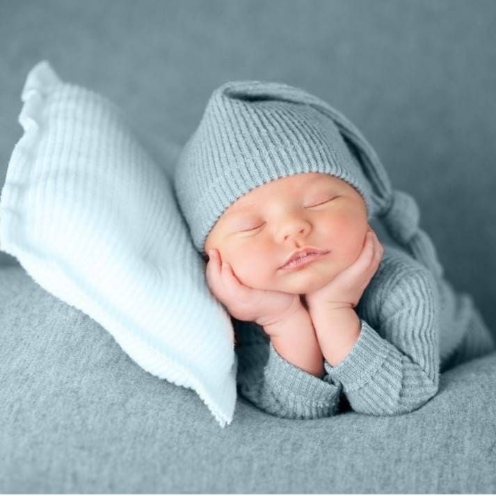 a sleeping baby wearing a blue hat on top of a gray blanket with white pillows