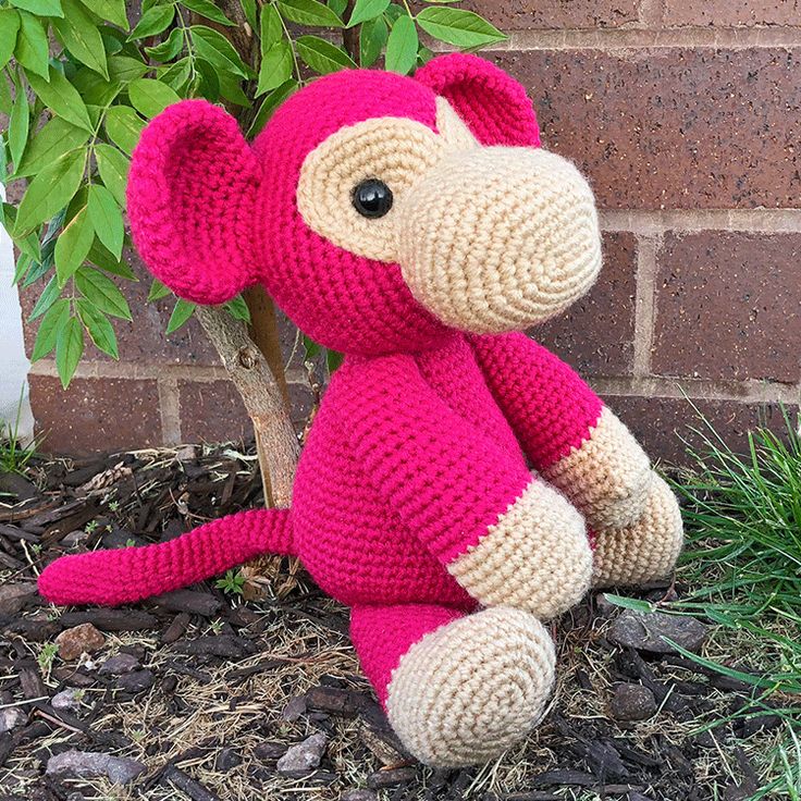 a crocheted pink and beige monkey sitting in front of a brick wall next to a tree