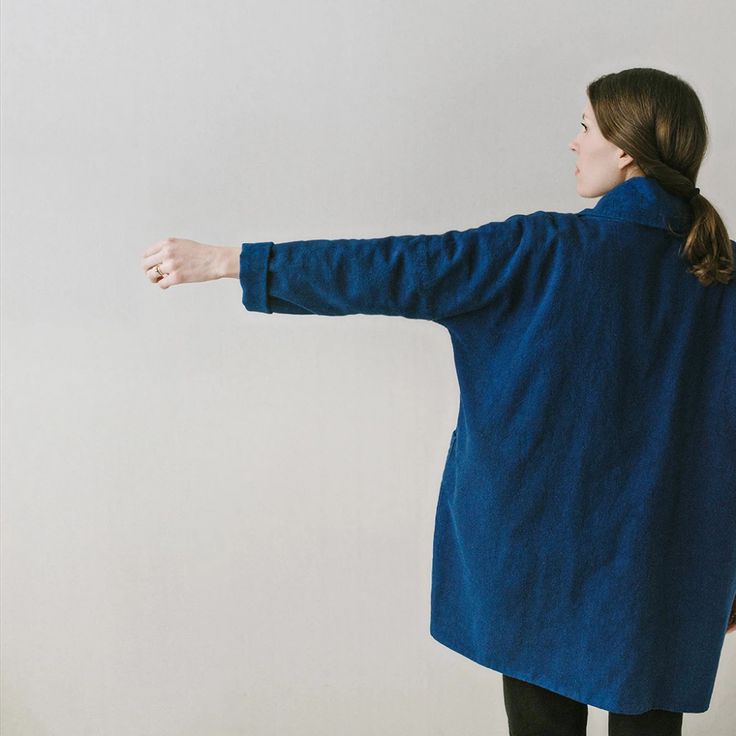 a woman standing with her arms outstretched in front of a white wall wearing a blue coat