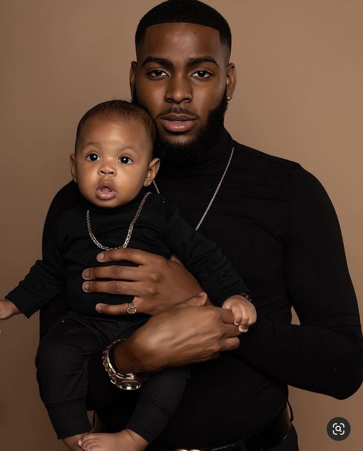 a man holding a baby in his arms and wearing a black turtle neck sweater with gold chains