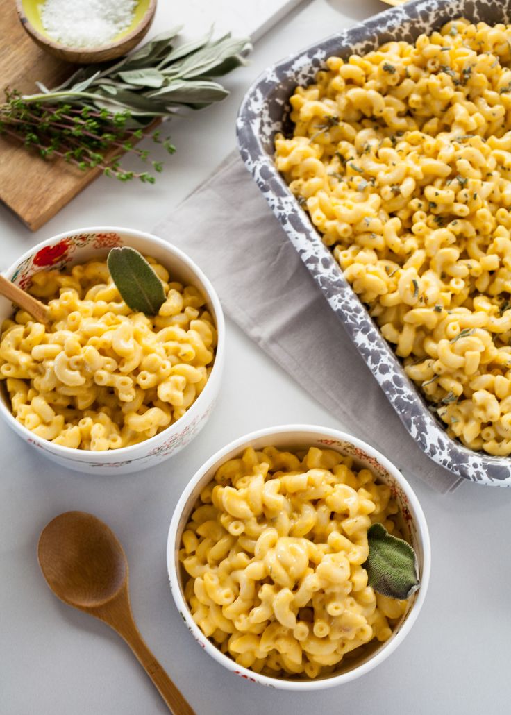 two bowls filled with macaroni and cheese on top of a white table next to wooden spoons