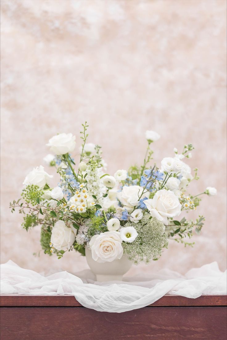 a vase filled with white and blue flowers on top of a wooden table next to a wall