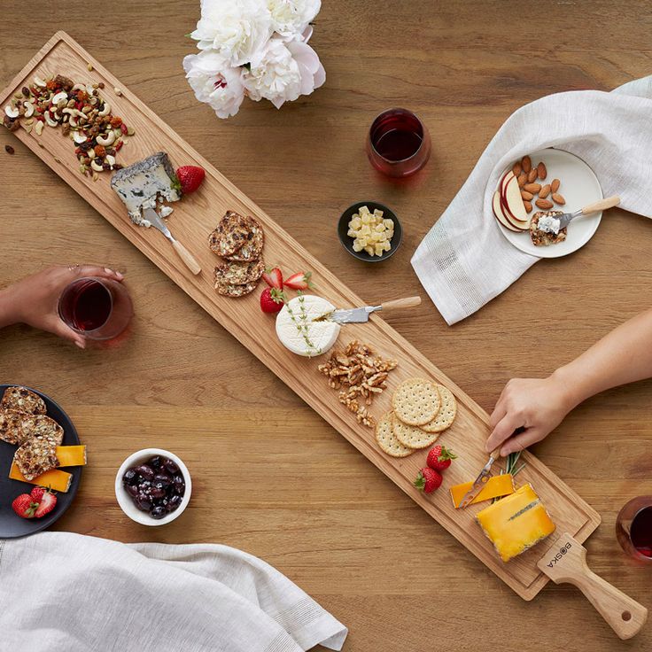 two people are serving themselves food on a long wooden board with cheeses and crackers